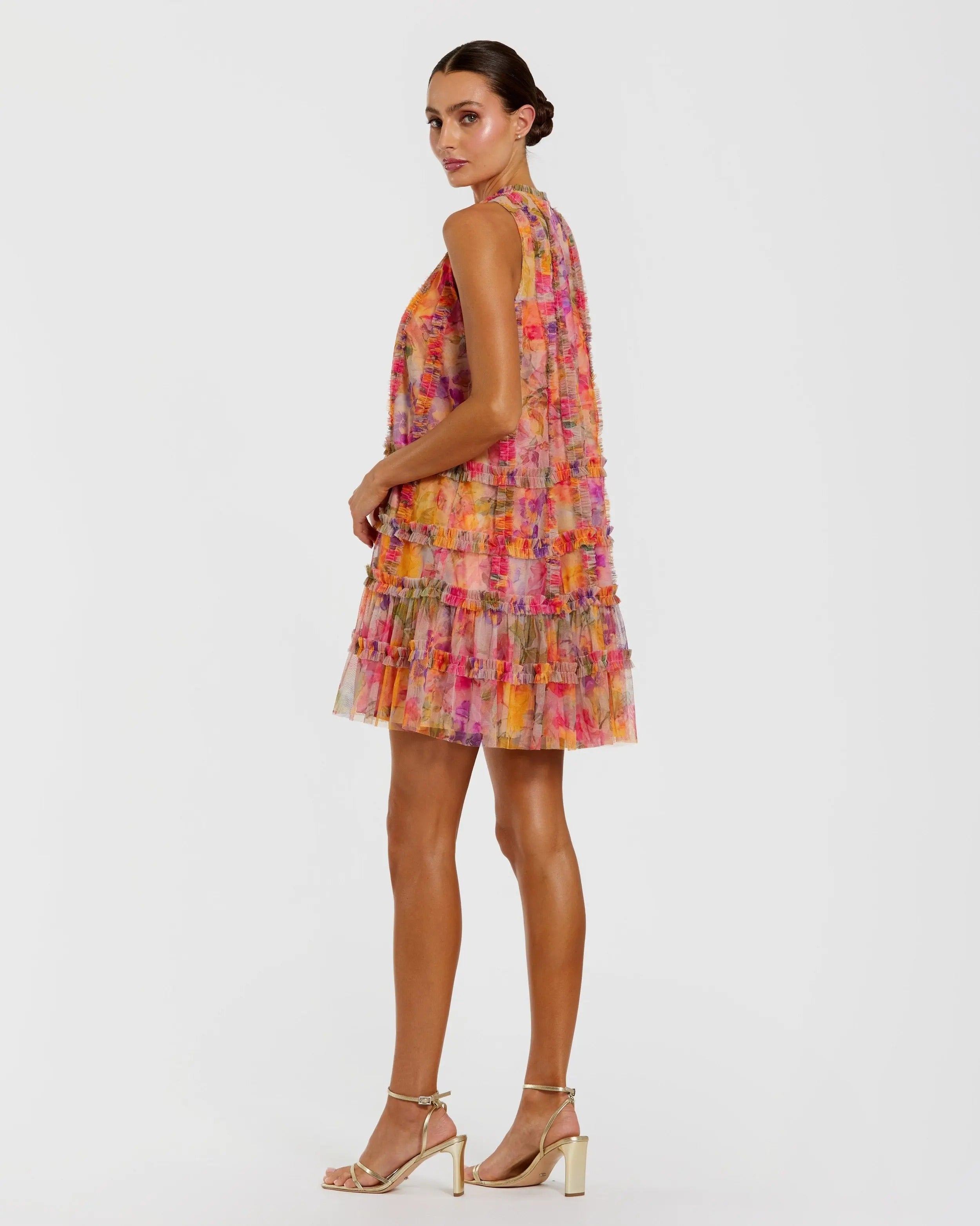 A woman standing in profile models the Mac Duggal 8488 short party dress, a sleeveless mini with a colorful floral pattern and ruffled, tiered layers, paired with gold high-heeled sandals against a plain white background.